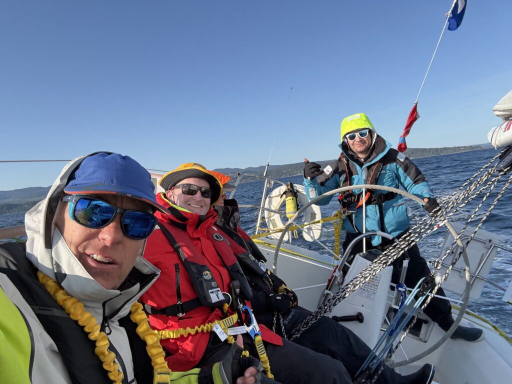 Three sailors on sailboat, one at helm, 2 sitting on lazarette, sailing in the straits of juan de fuca
