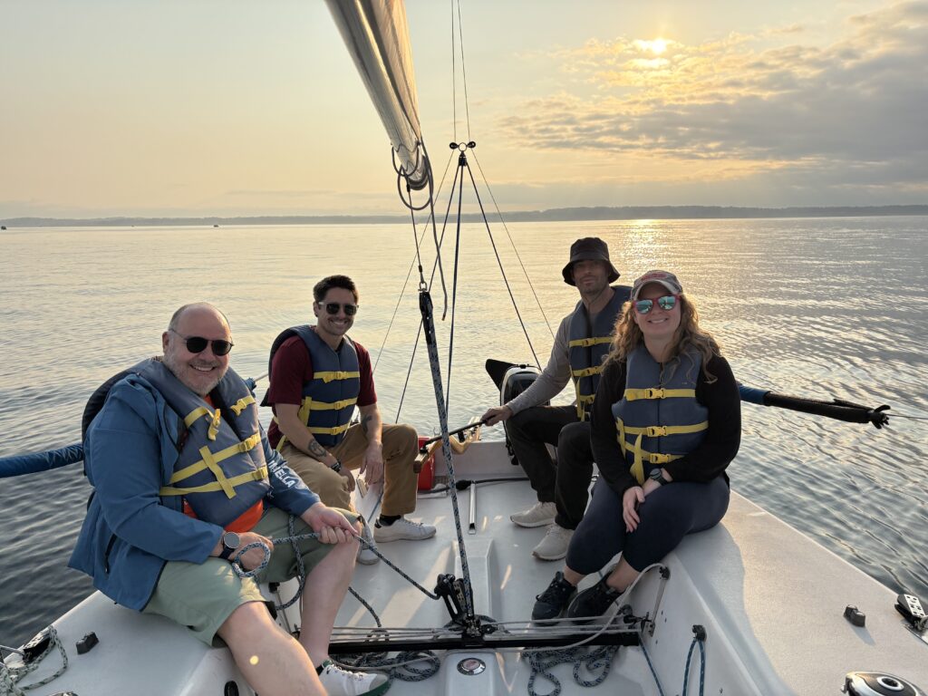 Four smiling people sitting on a sailboat on the water, with the sun setting behind them.